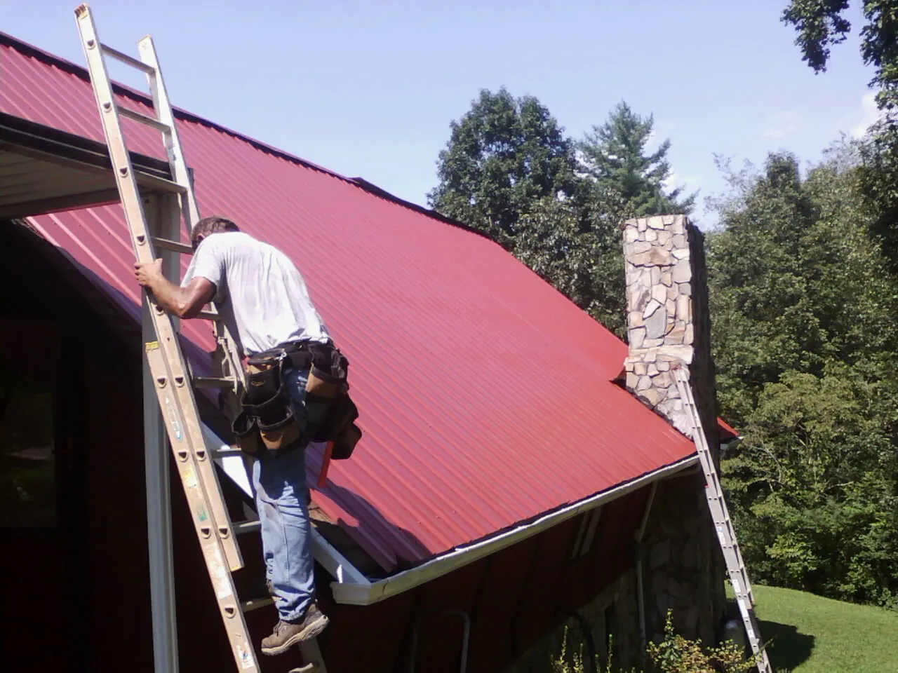 Roofer on a ladder beside a red metal roof and stone chimney — Soffit & Fascia Repair by Roofing Brothers in Columbia City